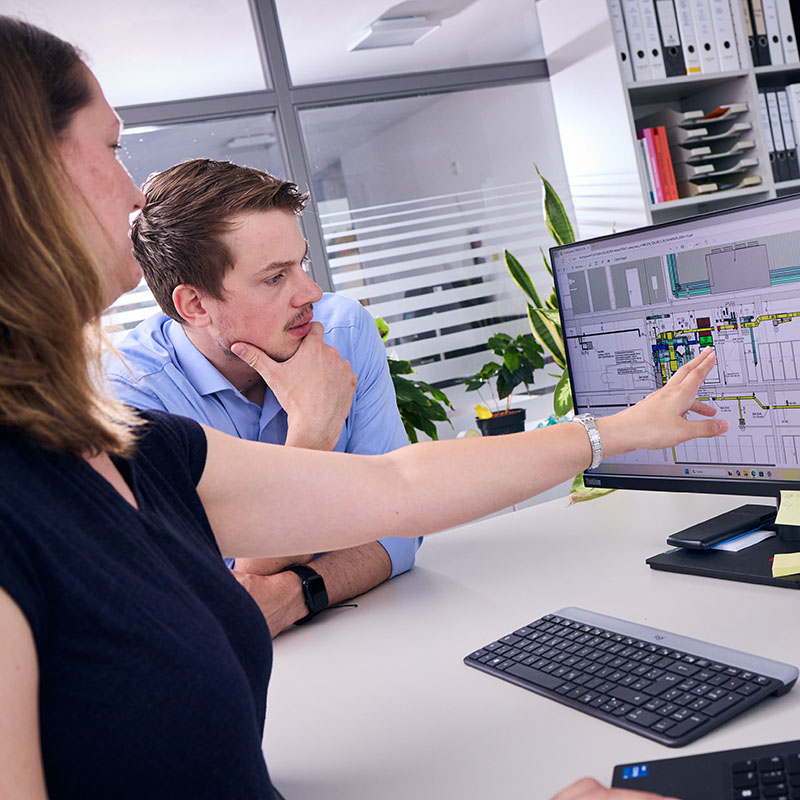 Zwei Mitarbeitende besprechen eine Bauzeichnung vor einem Monitor